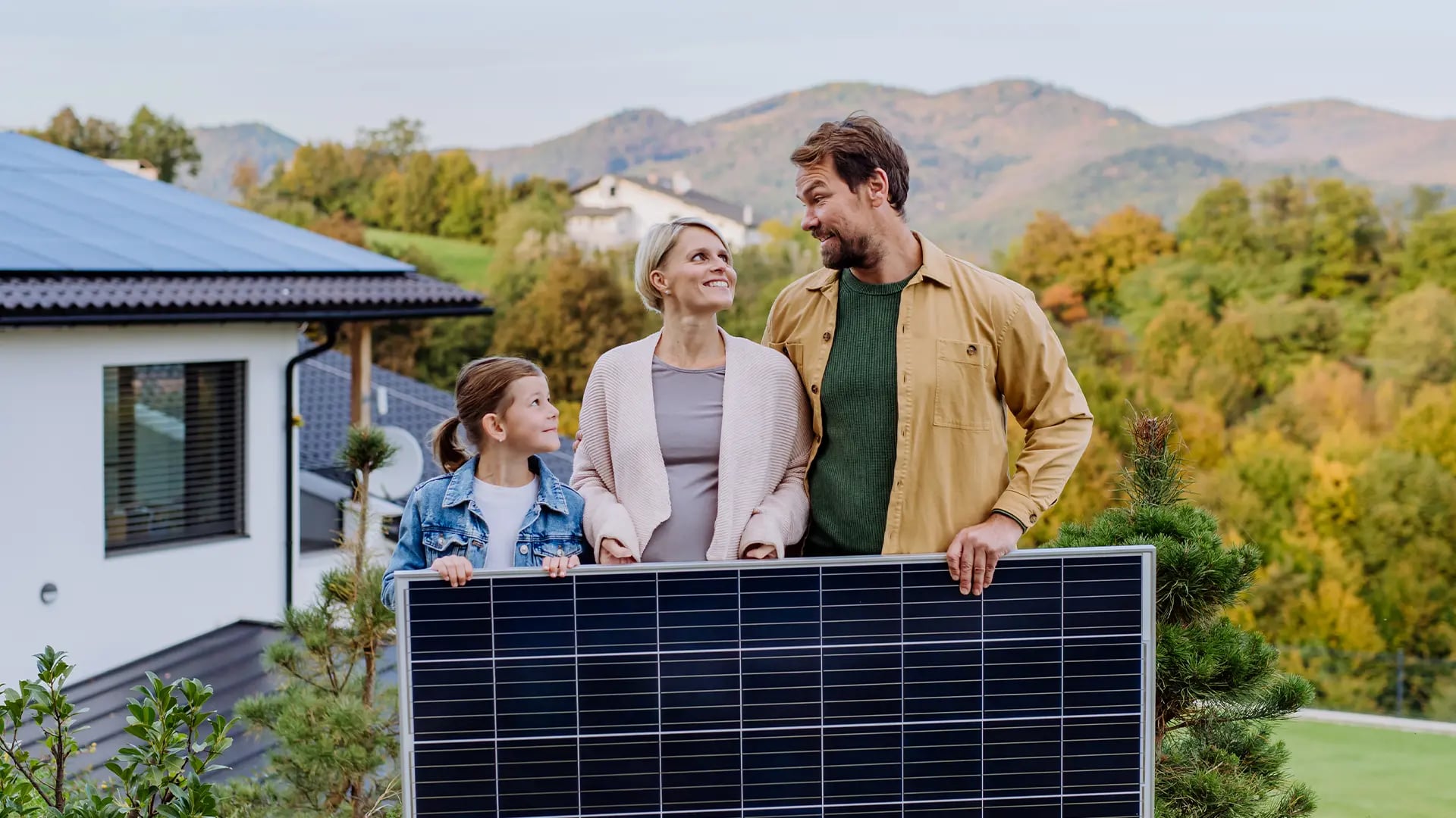 Familie mit Kind steht hinter einem Solarmodul im Garten, dahinter ein Wohnhaus mit Solardach