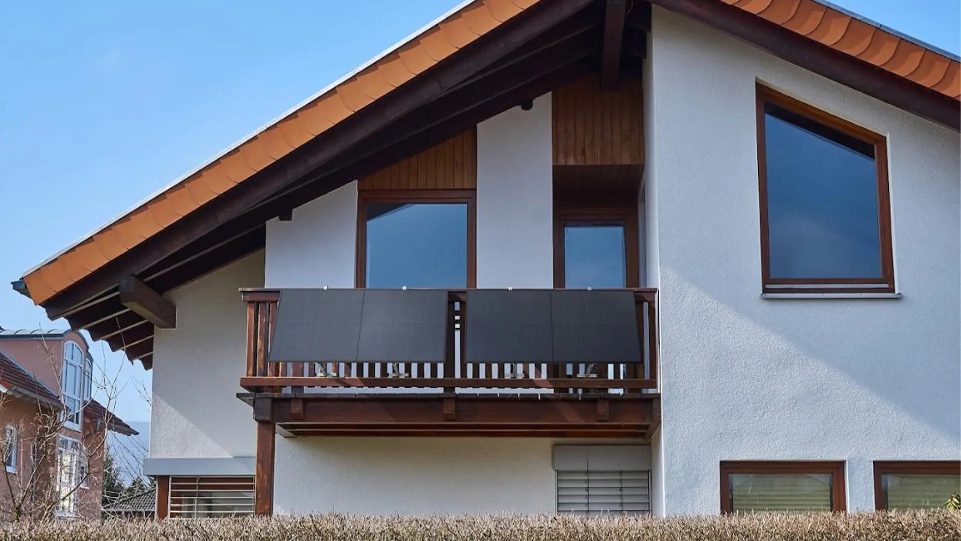 Zwei dunkle Solarmodule an der Holzbrüstung eines Balkons an einem Haus mit Ziegeldach