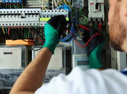 Elektriker mit grünen Schutzhandschuhen arbeitet an einem geöffneten Verteilerschrank mit Reihenklemmen und Sicherungsautomaten