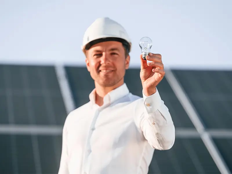 Mann mit weißem Schutzhelm und hellem Hemd hält eine Glühbirne in die Kamera, dahinter eine Reihe aufgeständerter Solarmodule