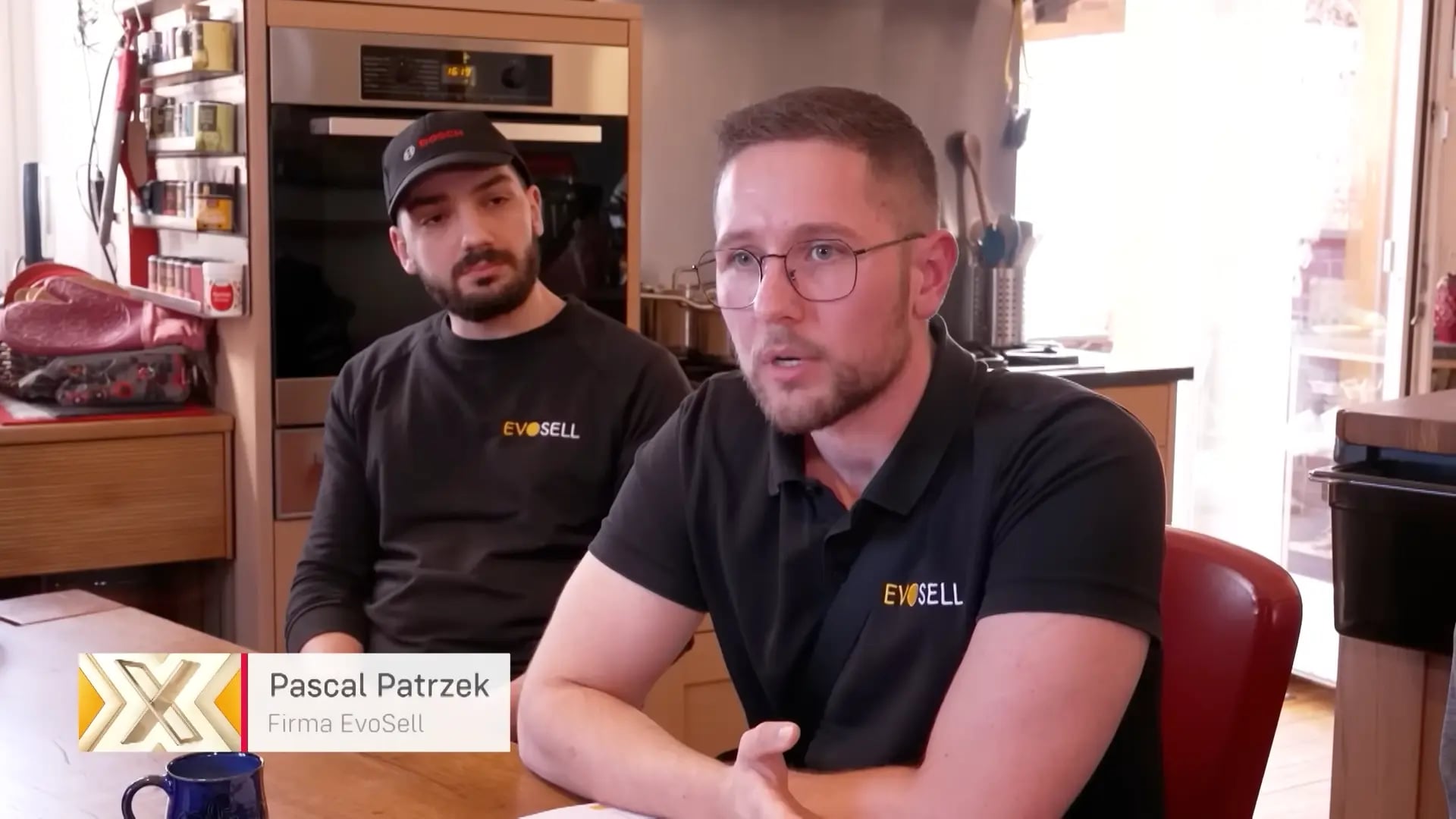 Standbild aus dem RTL+-Beitrag: zwei EvoSell-Mitarbeiter in schwarzen Firmenshirts sitzen in einer Küche im Gespräch, eingeblendet die Bauchbinde „Pascal Patrzek, Firma EvoSell“