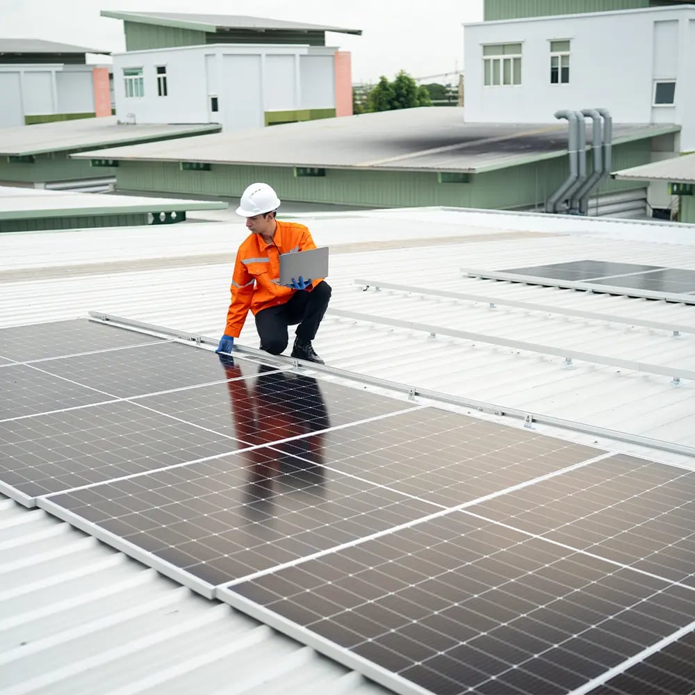 Monteur mit Helm und Laptop kniet auf einem flachen Blechdach neben montierten Solarmodulen
