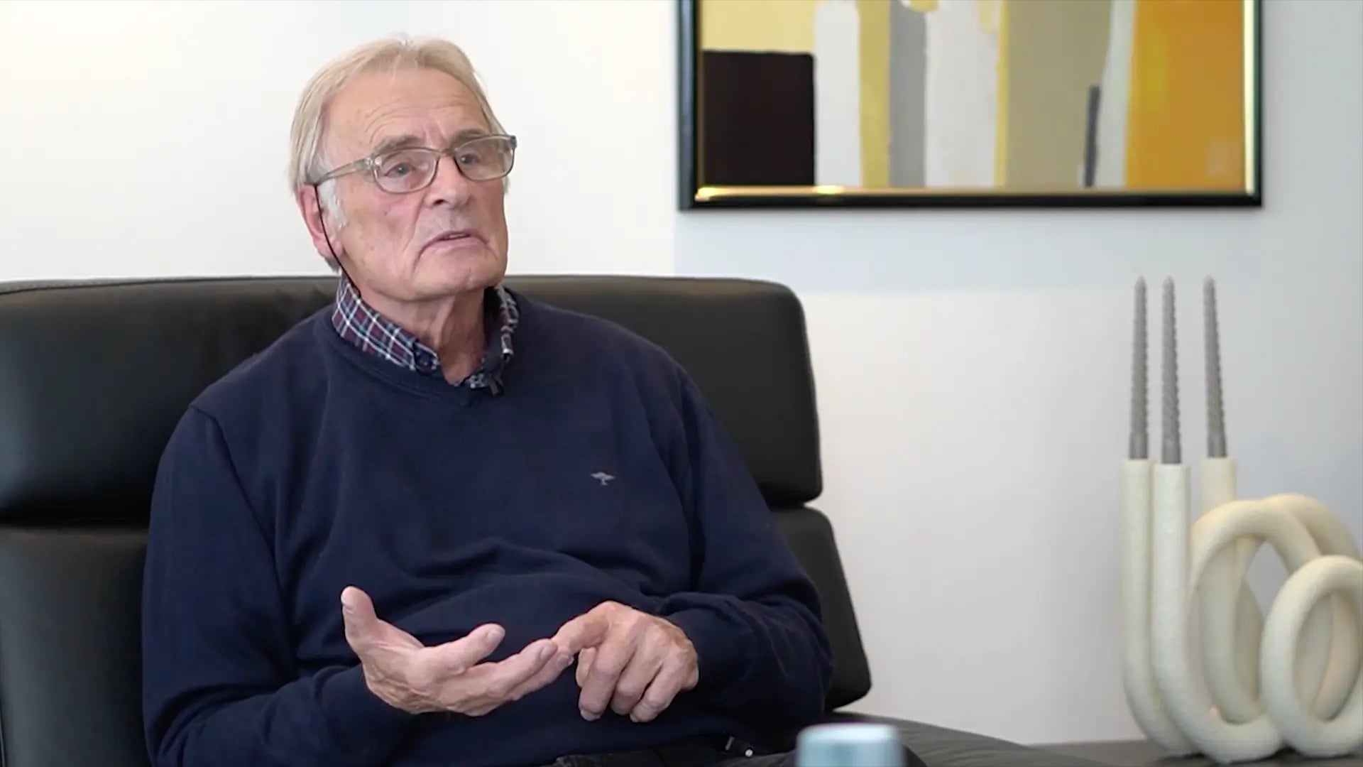 Standbild aus dem Kundenvideo: ein älterer Herr im dunkelblauen Pullover sitzt auf einem Ledersofa und erzählt, im Hintergrund ein Bild in Gelbtönen