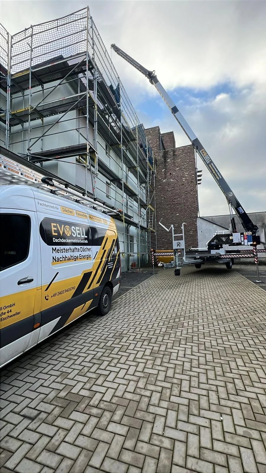 EvoSell-Transporter mit Dachdeckermeisterbetrieb-Beschriftung vor einem eingerüsteten Mehrfamilienhaus, rechts ein aufgestellter Kranausleger mit EvoSell-Logo