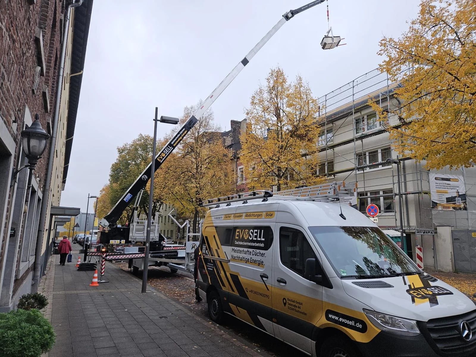 EvoSell-Transporter am Straßenrand vor einem eingerüsteten Haus, ein Autokran hebt Material aufs Dach, herbstlich gelbe Bäume